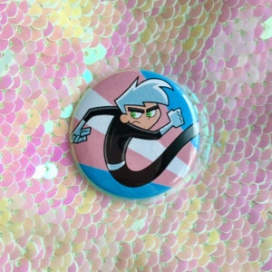 May include: A round button featuring a cartoon image of a character from the show Danny Phantom. The character is running and has a blue, pink and white transgender pride flag behind them.