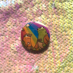 May include: A round button with a rainbow background and two frogs. One frog is green and wearing a brown jacket. The other frog is wearing a blue jacket. The text "Love is Love" is written on the button.
