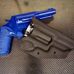 K&ouml;nnte beinhalten: Eine blaue Revolverpistole mit einem schwarzen Lederholster. Die Waffe hat einen strukturierten Griff und einen silbernen Lauf. Das Holster hat zwei Metalldruckkn&ouml;pfe.