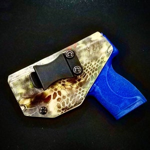 Puede incluir: Una funda de camuflaje con un clip negro y una pistola de plástico azul. La funda tiene una superficie texturizada y un clip de metal.