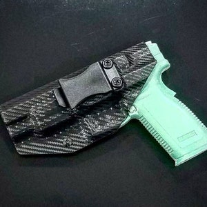 Op de afbeelding: Een zwarte carbonfiber holster met een zwarte clip en een lichtgroen pistool. De holster heeft een getextureerd oppervlak en is ontworpen om te worden gedragen op een riem.