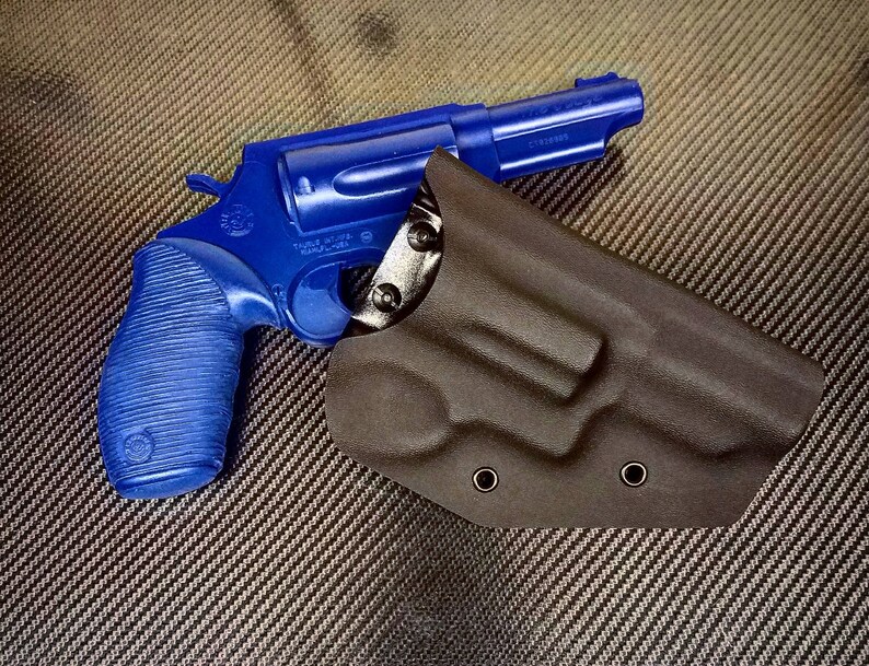 K&ouml;nnte beinhalten: Eine blaue Revolverpistole mit einem schwarzen Lederholster. Die Waffe hat einen strukturierten Griff und einen silbernen Lauf. Das Holster hat zwei Metalldruckkn&ouml;pfe.