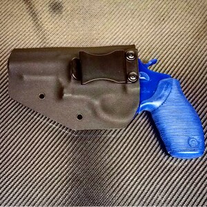 K&ouml;nnte beinhalten: Ein schwarzes Lederholster mit einem blauen Revolver darin. Das Holster hat einen Metallclip an der Seite.