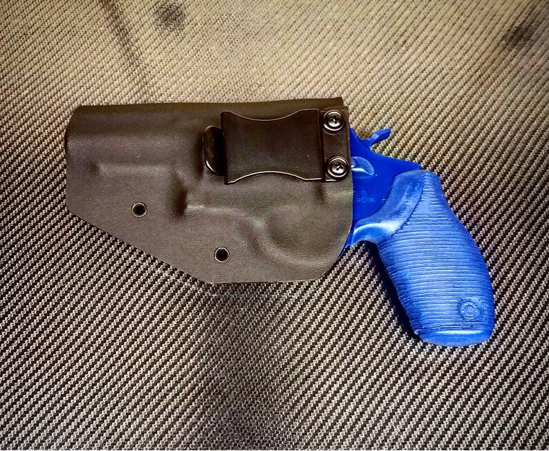 K&ouml;nnte beinhalten: Ein schwarzes Lederholster mit einem blauen Revolver darin. Das Holster hat einen Metallclip an der Seite.
