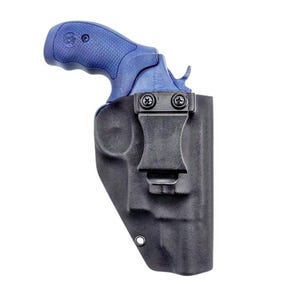 Op de afbeelding: Een blauwe revolver met een getextureerde grip is gedeeltelijk in een zwarte, gevormde holster geplaatst. De holster is voorzien van een zwarte metalen riemclip die met twee schroeven is bevestigd en een metalen oogje aan de onderrand.