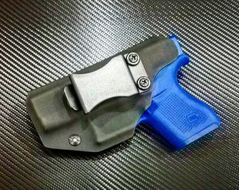 Puede incluir: Una funda de cuero negra con un clip de metal para una pistola azul. La funda tiene una superficie texturizada y un pequeño tornillo en el lateral.