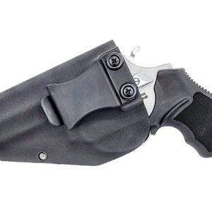 Può includere: Un fodero in pelle nera con una clip per trasportare un piccolo revolver argentato. Il revolver ha un'impugnatura nera e una canna argentata.