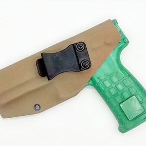 Puede incluir: Una funda de cuero marrón con un clip negro para una pistola. La funda está diseñada para llevarse en un cinturón. La pistola es verde y tiene un agarre texturizado.