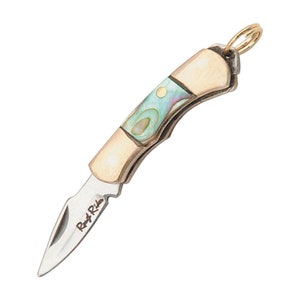 Puede incluir: Un pequeño cuchillo de bolsillo de tono dorado con un mango de nácar y una hoja plateada. La hoja está grabada con las palabras "Roy & Ride".