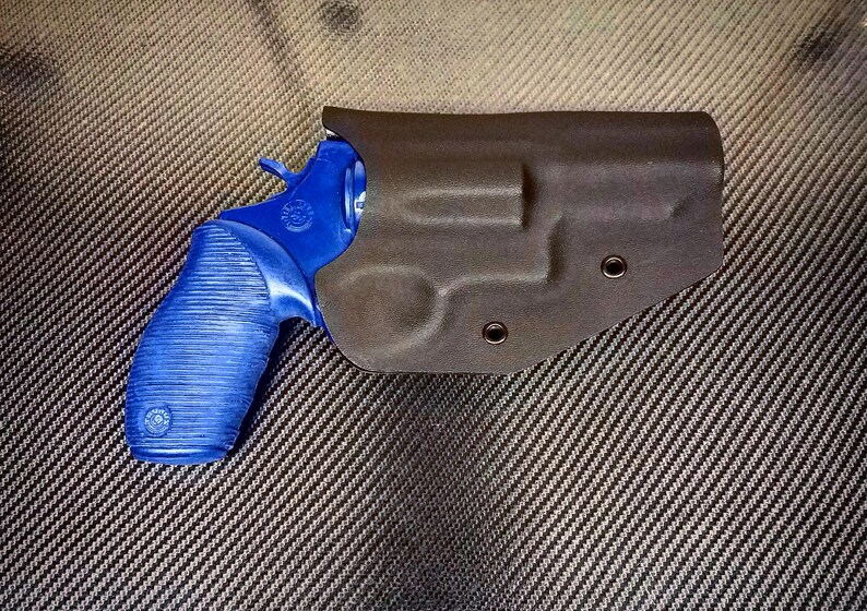 K&ouml;nnte beinhalten: Ein blauer Revolver mit einem schwarzen Lederholster. Das Holster hat zwei Druckkn&ouml;pfe.