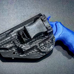 Op de afbeelding: Een holster met een zwart koolstofvezelpatroon en een zwarte metalen riemclip, met daarin een felblauwe revolver met een getextureerde grip. De holster heeft een gevormde pasvorm voor het vuurwapen, met zichtbare schroeven die de clip bevestigen.