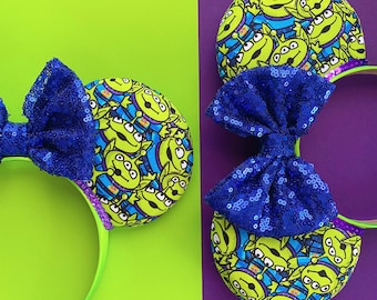 Puede incluir: Dos diademas con grandes lazos azules con lentejuelas y orejas redondas. Las orejas están cubiertas con un patrón de alienígenas verdes sobre un fondo azul. Las diademas son verdes y el fondo está dividido entre verde lima y morado.