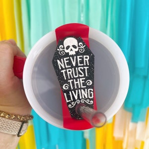Puede incluir: Un vaso rojo y blanco con una etiqueta en forma de lápida negra y blanca con un gráfico de calavera en blanco y negro y el texto "Never Trust The Living".