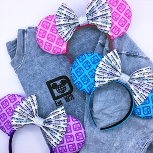May include: Several colorful mouse ear headbands with patterned ears and bows, resting on a gray t-shirt. The ears feature a repeating globe design in pink, blue, and purple. The bows have a train pattern with glitter accents. The t-shirt has a black logo that says "Est. 1971".
