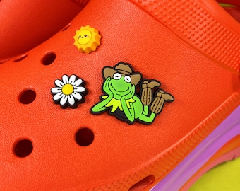 May include: Bright orange clogs with decorative charms. Charms include a smiling yellow sun, a white daisy with a yellow center, and a green frog wearing a cowboy hat. The clogs have a purple and pink sole.