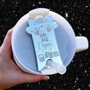 May include: A silver-colored metal tumbler lid with a UFO design and the text "Now I'm down bad, cryin' at the gym".