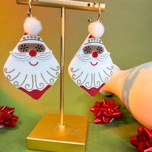 May include: A pair of festive Santa Claus earrings. Each earring features a white face with a red nose and beard, a brown mustache, and a red and white hat with a white pom-pom. The earrings are displayed on a gold stand.