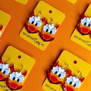 May include: Earrings featuring a cartoon duck design with red devil horns, white and blue eyes, and a red collar. The earrings are on yellow cards with the text "BEST DAY EVER Ears co." on an orange background. The earrings have gold-colored hoops.