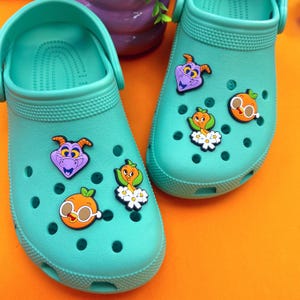 May include: Two pairs of teal blue clogs with various cartoon character shoe charms. The charms include a purple dragon, an orange bird with flowers, and an orange with sunglasses.