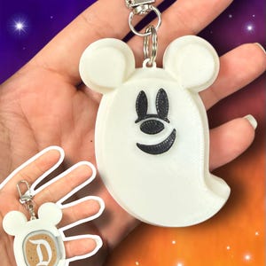 May include: A white ghost-shaped keychain with mouse-like ears and a black face featuring oval eyes and a curved mouth. The keychain includes a silver metal lobster clasp and ring. A smaller inset shows the reverse side with a coffee cup graphic and letter D.