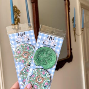 May include: Two sets of circular car coasters in clear plastic packaging with blue and white checkered backing cards labeled CAR COASTERS. One set features a green leaf pattern, while the other displays a colorful floral and butterfly design.
