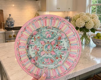 May include: A round ceramic plate featuring a decorative border with alternating pink and light green stripes. The center displays a circular medallion surrounded by four fan-shaped panels, each decorated with intricate illustrations of figures and flowers.
