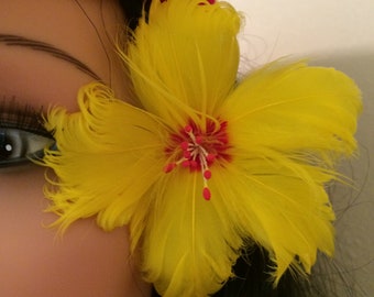 Può includere: Un fermaglio per capelli a forma di fiore giallo di piume con un centro rosso. Il fiore è sulla testa di una persona, vicino al suo occhio.