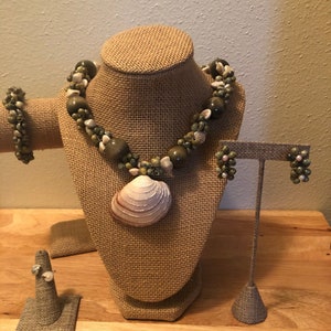 Könnte beinhalten: Ein Set aus grünem und braunem Muschelschmuck, bestehend aus einer Halskette, einem Armband und Ohrringen. Die Halskette hat einen großen Muschel-Anhänger. Der Schmuck wird auf einer Jute-Puppe und einem Ständer präsentiert.
