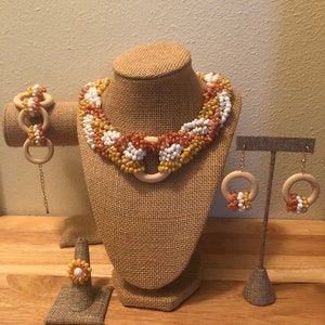 Könnte beinhalten: Ein Schmuckset mit einer Halskette, einem Armband, Ohrringen und einem Ring. Der Schmuck ist aus Holz und weißen, orangefarbenen und gelben Perlen gefertigt. Die Halskette und die Ohrringe haben die Form eines Kreises.