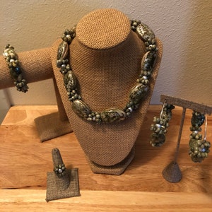 Könnte beinhalten: Ein Set aus grünem und weißem Muschelschmuck, bestehend aus einer Halskette, einem Armband, einem Ring und Ohrringen. Der Schmuck wird auf einer mit Sackleinen bezogenen Puppe und Ständern präsentiert.