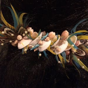 Puede incluir: Un peine para el cabello con una fila de conchas y plumas. Las conchas son blancas y tienen una textura natural. Las plumas son azules, amarillas y marrones.