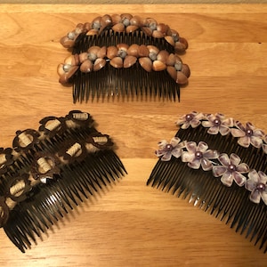 Puede incluir: Tres peines decorativos para el cabello con diferentes diseños. Uno presenta pequeñas conchas de color marrón claro, otro tiene adornos en forma de flor de color marrón oscuro con centros beige, y el tercero tiene flores moradas y blancas con cuentas moradas.