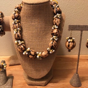 Könnte beinhalten: Ein Schmuckset mit Halskette, Armband, Ring und Ohrringen. Die Halskette und das Armband bestehen aus großen, oval geformten Perlen mit braunen und beigen Mustern, die mit kleineren weißen, gelben und grünen Perlen durchsetzt sind. Die Ohrringe und der Ring haben ein ähnliches Design.