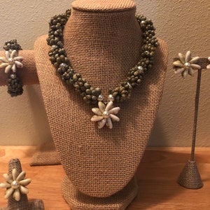 Könnte beinhalten: Ein Set aus grünem und weißem Muschelschmuck, bestehend aus einer Halskette, einem Armband und Ohrringen. Die Halskette hat einen großen Blumenanhänger.