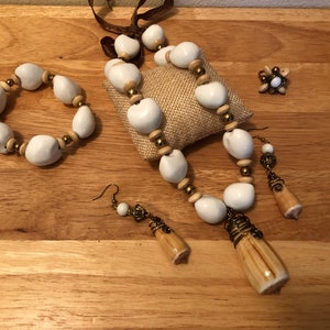 Puede incluir: Un conjunto de collar, pulsera y pendientes de cuentas de semillas blancas y marrones. El collar tiene un largo colgante hecho de una vaina de semillas. El conjunto se muestra sobre una superficie de madera.