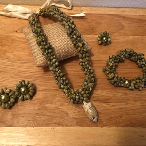 Könnte beinhalten: Ein komplettes Schmuckset mit Halskette, Armband, Ohrringen und einem Ring. Das Set besteht aus kleinen, olivgrünen Perlen und einem Anhänger. Die Ohrringe und der Ring haben eine goldfarbene Mitte. Der Schmuck wird auf einer Holzoberfläche präsentiert.
