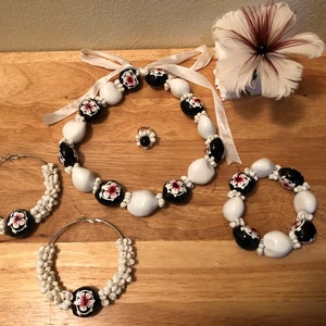 以下が含まれることがあります： ネックレス、ブレスレット、イヤリングを含む、白と黒の花柄のジュエリーセット。ジュエリーは、黒と赤の花のアクセントが付いた白いビーズで作られています。
