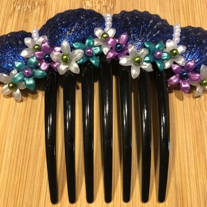 Puede incluir: Un peine decorativo para el cabello con un diseño de concha marina azul oscuro y brillante. El peine está adornado con pequeñas flores de colores blanco, turquesa y morado, con detalles de perlas. Los dientes del peine son negros.