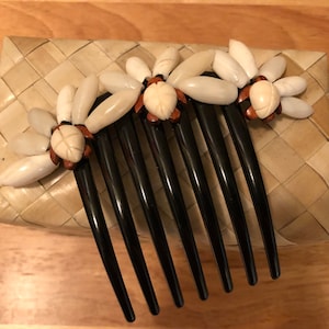 Puede incluir: Un peine de plástico negro para el cabello con tres acentos de flores de concha blanca. Cada flor tiene un amuleto de tortuga marrón en el centro.