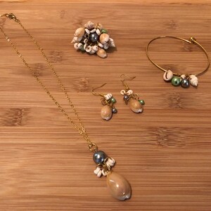 May include: A set of gold-tone jewellery featuring a necklace, bracelet, and earrings. Each piece is adorned with small seashells, pearls, and beads in shades of white, green, and grey.