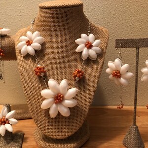 Könnte beinhalten: Ein Schmuckset aus einer weißen Muschelblume Halskette, Armband, Ring und Ohrringen. Die Blumen sind aus weißen Muscheln mit orangefarbenen Zentren. Der Schmuck befindet sich auf einem weißen Hintergrund.