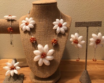 Könnte beinhalten: Ein Schmuckset aus einer weißen Muschelblume Halskette, Armband, Ring und Ohrringen. Die Blumen sind aus weißen Muscheln mit orangefarbenen Zentren. Der Schmuck befindet sich auf einem weißen Hintergrund.