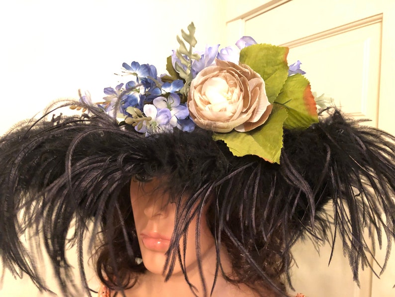 Pu&ograve; includere: Un cappello nero a larghe falde con un grande pennacchio di piume nere. Il cappello &egrave; adornato da un gruppo di fiori blu e bianchi e da un unico grande fiore beige.