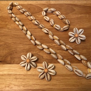 Puede incluir: Un conjunto de joyas de conchas que incluye un collar largo, una pulsera y dos pares de pendientes. El collar y la pulsera están hechos de pequeñas conchas blancas, y los pendientes tienen forma de flores con un pequeño centro naranja.