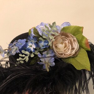 Pu&ograve; includere: Un cappello nero con una grande piuma di piume nere e un'arrangiamento floreale in cima. L'arrangiamento include fiori blu e bianchi, un grande fiore beige e foglie verdi.