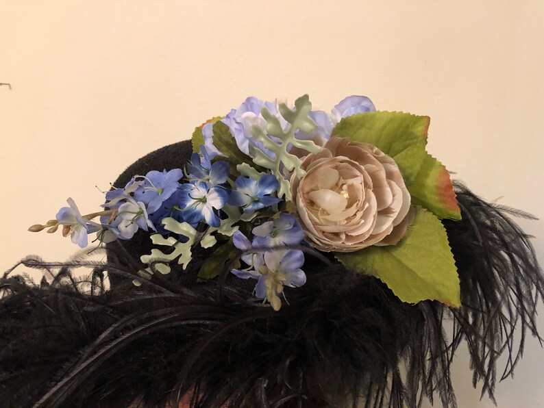 Pu&ograve; includere: Un cappello nero con una grande piuma di piume nere e un'arrangiamento floreale in cima. L'arrangiamento include fiori blu e bianchi, un grande fiore beige e foglie verdi.
