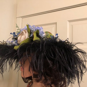 Pu&ograve; includere: Un cappello nero a tesa larga con un grande pennacchio di piume nere. Il cappello &egrave; decorato con un gruppo di fiori artificiali bianchi e blu.