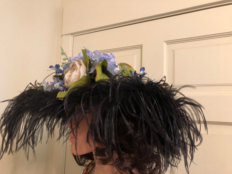 Pu&ograve; includere: Un cappello nero a tesa larga con un grande pennacchio di piume nere. Il cappello &egrave; decorato con un gruppo di fiori artificiali bianchi e blu.