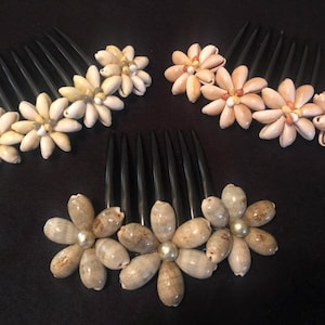 May include: Three hair combs with decorative flower designs made from seashells. The combs are black plastic with a row of teeth. The flowers are made from different types of seashells in shades of white, beige, and brown.