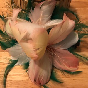 Op de afbeelding: Een bloemen haarclip met gelaagde veren in roze, wit en geel, geaccentueerd met groene veren. De clip is ontworpen om op een bloem te lijken, perfect om een vleugje elegantie aan elk kapsel toe te voegen.
